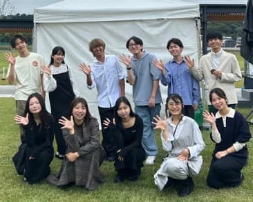 野外の白いテントの前で手を振るアカペラサークルの部員11人の集合写真