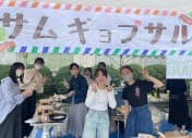 「サムギョプサル」の横断幕を掲げた屋台の前でポーズをとる部員たち