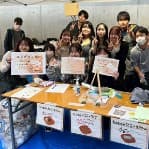 模擬店のテーブルの後ろでメニューの紙を持ってポーズをとる部員たち
