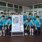 建物の入口前で看板を囲んで集合写真を撮る、水色のTシャツ姿の卓球部の部員たち