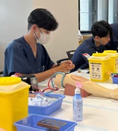 スクラブ姿の学生が腕の模型で採血の練習をしている写真